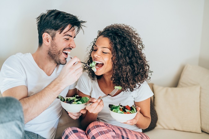 mixed man and woman on couch, laughing and eating healthy salad with the man feeding the woman