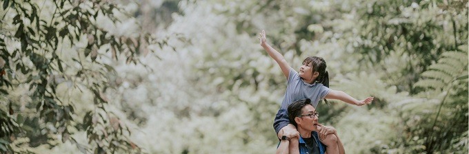 Asian father with daughter on his shoulders walking outside with lots of greenery around them
