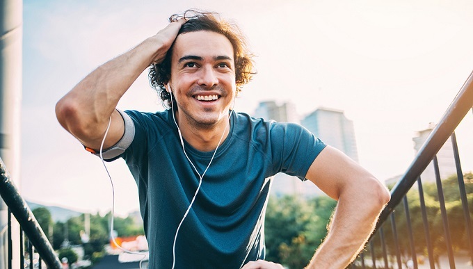 white young male listening to music with earbuds outside during the summer and smiling